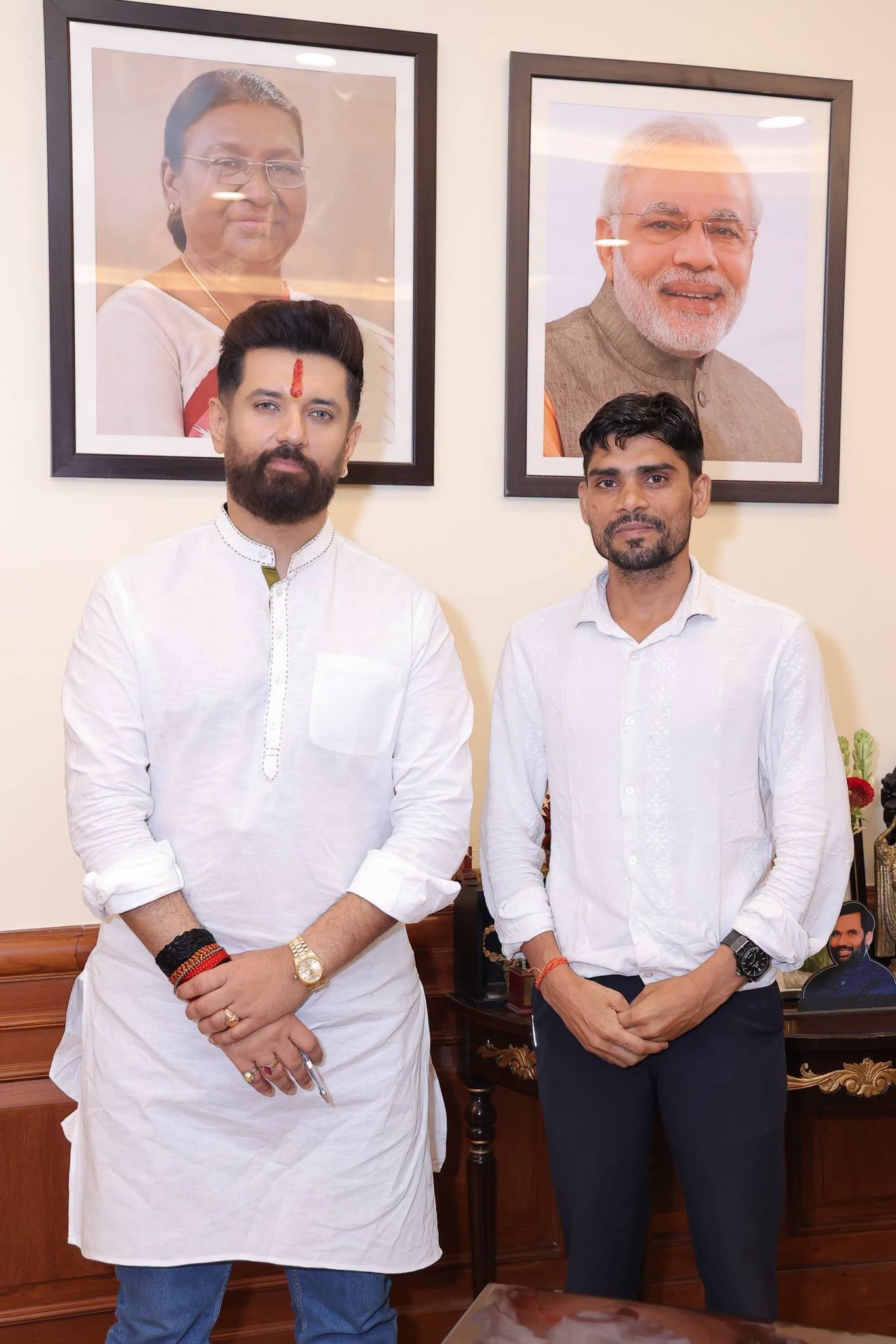Two men in white kurta and shirt standing side by side in an office, framed portraits of national leaders on the wall behind