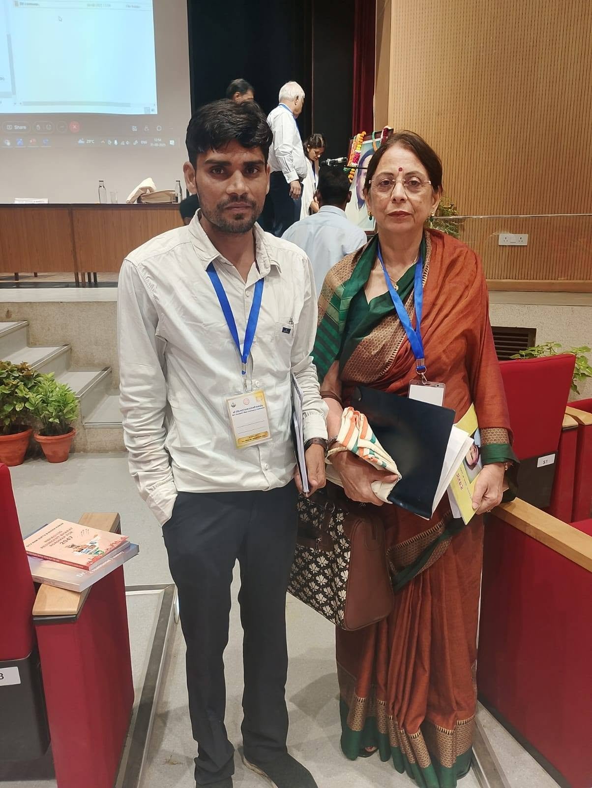 A man wearing a conference lanyard standing beside a woman in a saree inside an auditorium