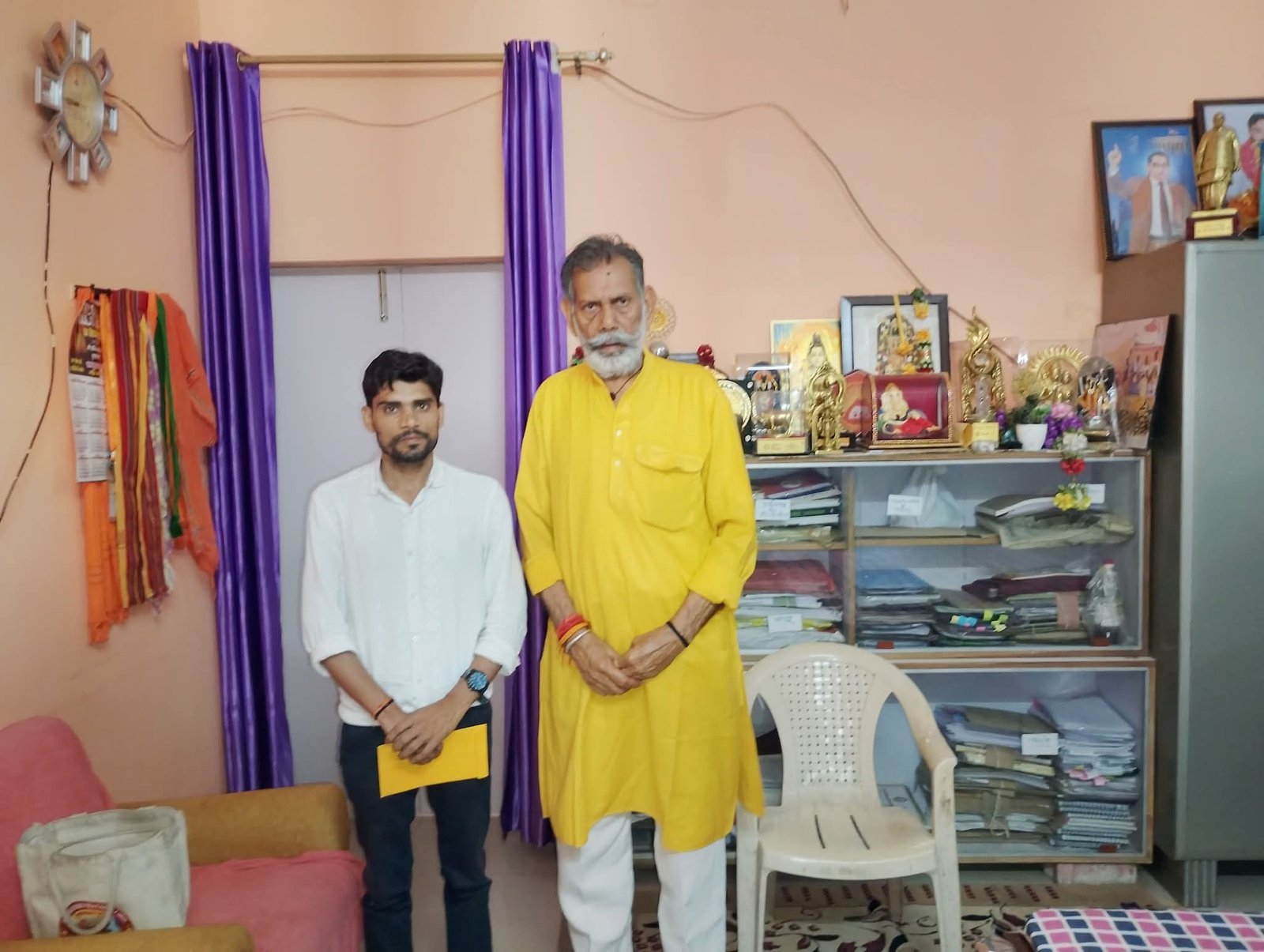 A man holding an envelope standing beside an older man in a yellow kurta inside a room with shelves and trophies