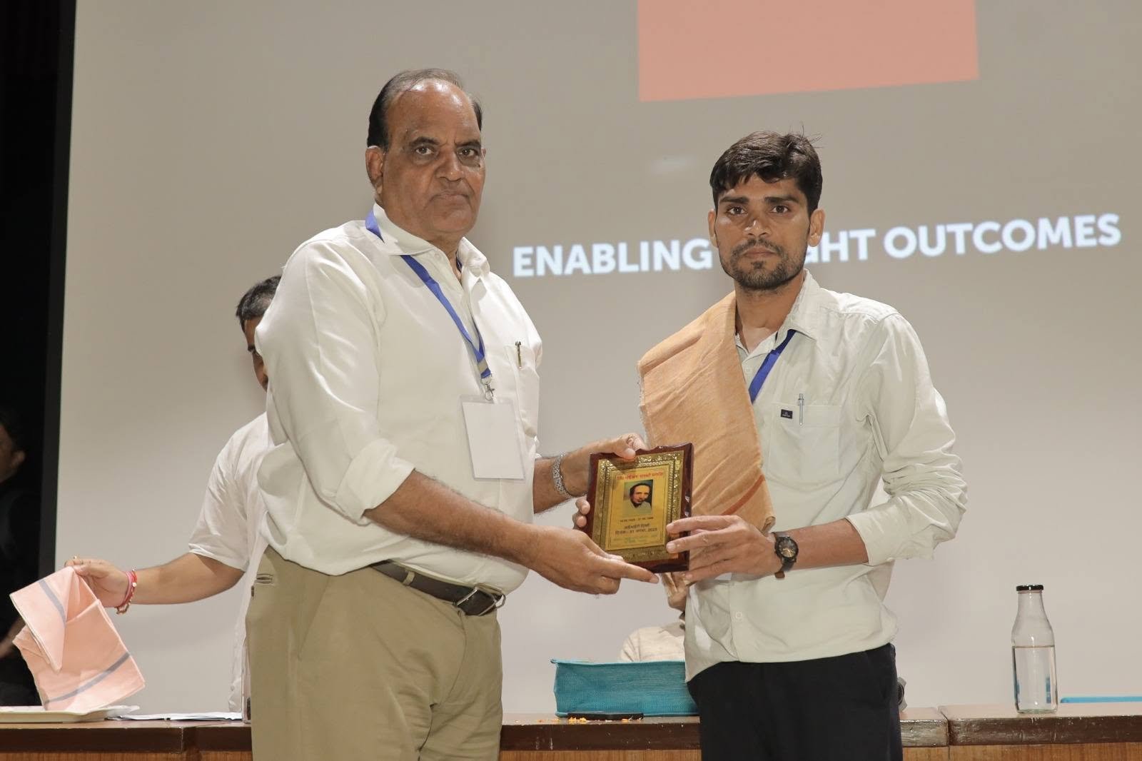 A man wearing a shawl receiving a framed memento from an older man on stage at an indoor event