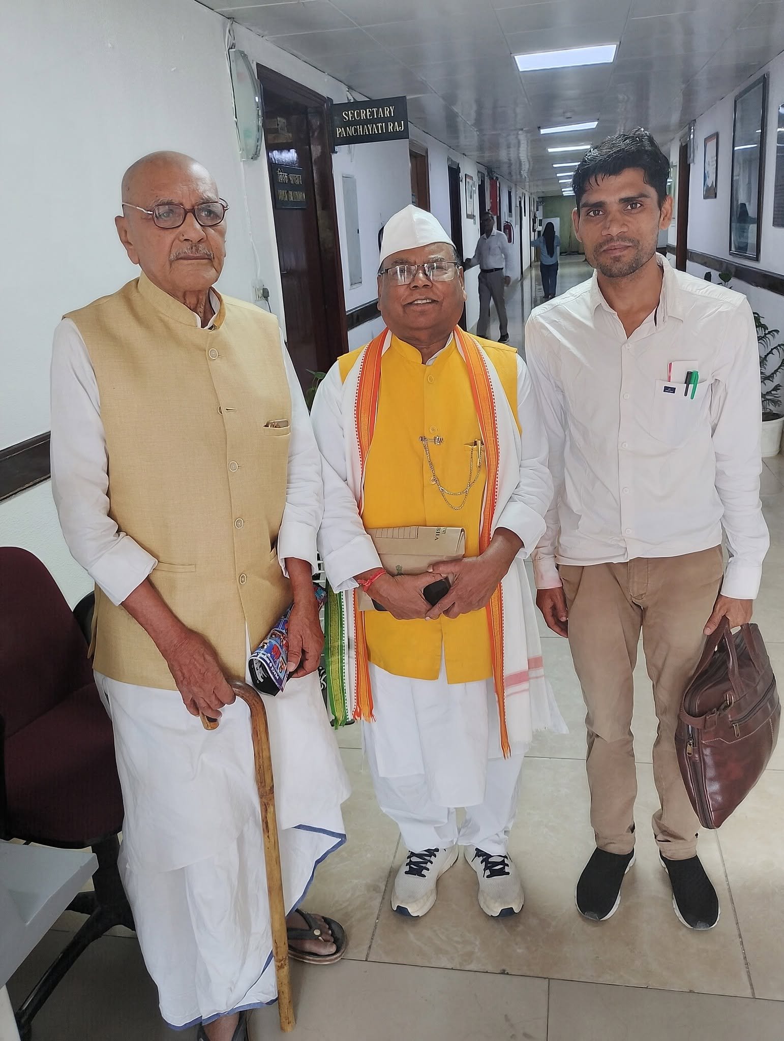 Three men standing in a government office corridor beneath a sign reading Secretary Panchayati Raj