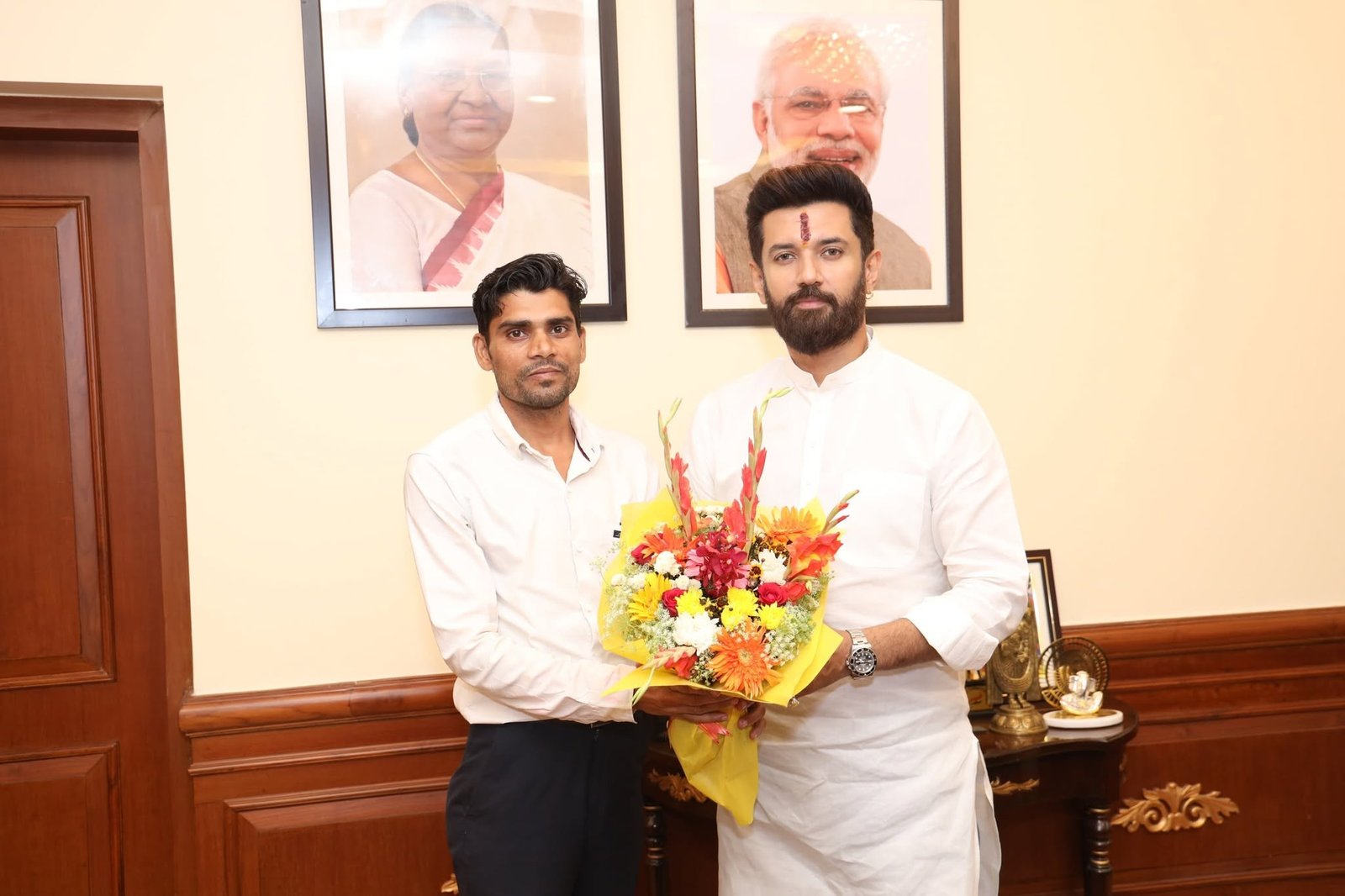 Two men holding a bouquet of flowers together in an office, framed portraits of national leaders on the wall behind