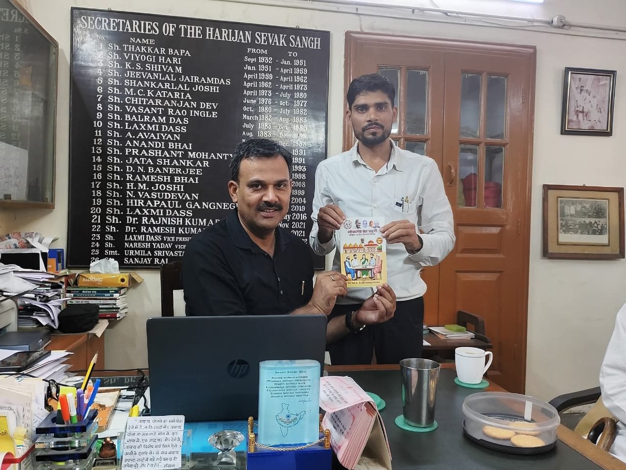 Two men holding an illustrated booklet in an office, an honours board listing past secretaries behind them