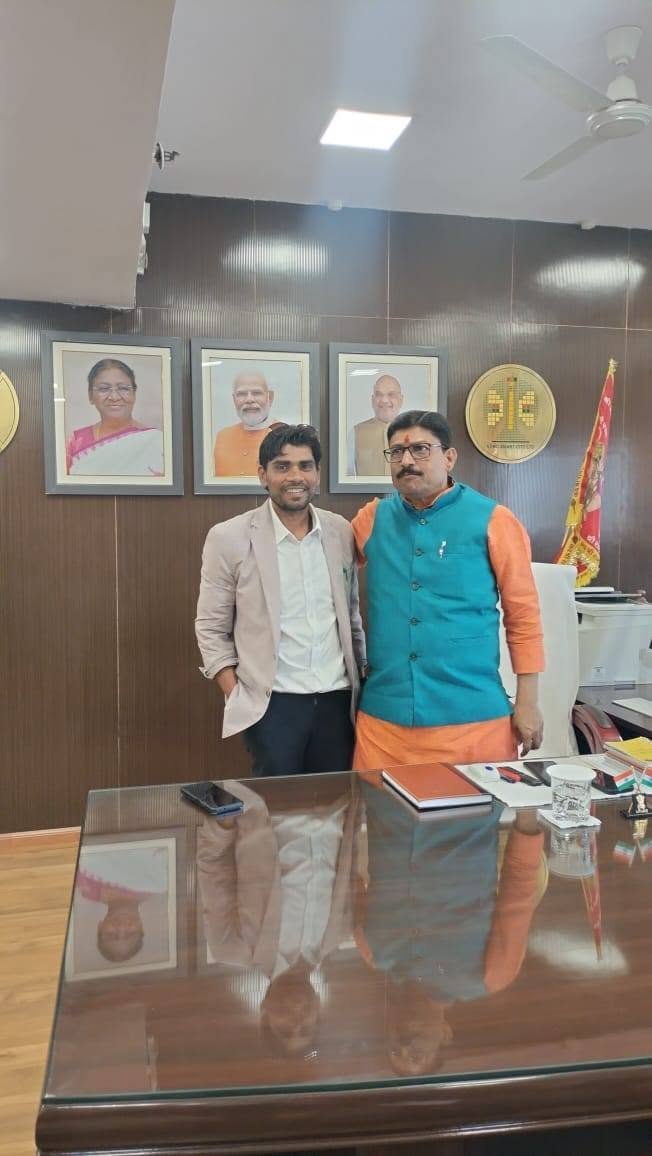 Two men standing behind a polished desk in an office, framed portraits of national leaders on the wall behind